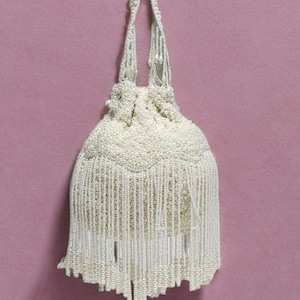 May include: An off-white beaded purse with a drawstring closure and long fringe. The purse is made of small, round beads and hangs against a pink background. The handle is also beaded.
