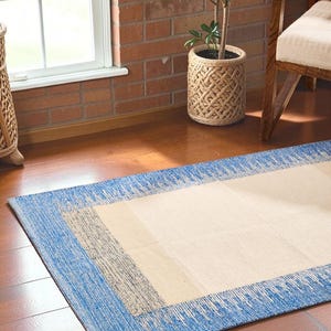 May include: A rectangular rug with a blue border and a beige centre. The rug has a textured appearance and is placed on a hardwood floor. A small potted plant in a woven basket is visible in the background.