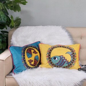 May include: Two decorative throw pillows with fish designs. One pillow is blue with a fish design, the other is yellow with a fish design. Both pillows have a pink border and are on a white furry blanket.
