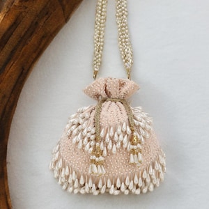 May include: A blush pink pouch bag with a pearl-like beaded handle. The bag is decorated with numerous white teardrop-shaped beads and gold tassels, creating an elegant design. The bag is a small, round shape, ideal for special occasions.