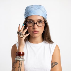 Puede incluir: Un pañuelo azul claro con estampado de cachemira azul oscuro sobre la cabeza, combinado con gafas ovaladas de carey. El conjunto incluye una pila de pulseras con un brazalete marrón jaspeado, cadenas plateadas y una banda verde claro lisa.