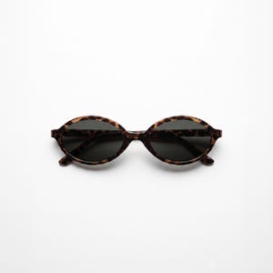 Peut inclure: Lunettes de soleil de forme ovale avec une monture au motif écaille de tortue marron et noir. Les verres sont sombres, d'un noir uni, et offrent une couverture complète à l'intérieur des montures arrondies. Le pont relie les deux verres.