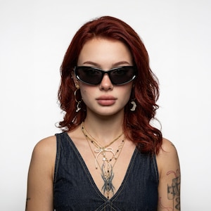 May include: Black oval-shaped sunglasses with dark lenses. The jewelry includes gold-toned hoop earrings with a crescent moon charm, a layered gold-toned necklace featuring a small white bow, black beads, and a star charm, worn over a dark denim top.