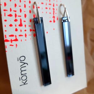 Könnte beinhalten: Ein Paar dunkelblaue, rechteckige Ohrringe mit silberfarbenen Haken. Die Ohrringe werden auf einer weißen Karte mit einem roten Punktmuster und dem Wort "kōmyō" in Schwarz präsentiert.