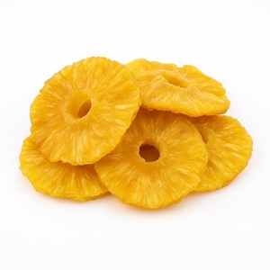 以下が含まれることがあります： 鮮やかな黄色が特徴の、乾燥パイナップルの輪切りの山。輪切りは中央に穴が開いた円形で、質感がはっきりと見て取れます。画像は無地の白い背景に配置されています。