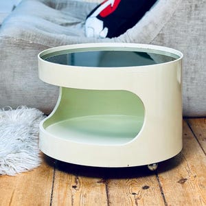 May include: A cream-colored, mid-century modern side table with a circular top and a rounded, open design. The table has a dark glass top and sits on small wheels. The table is in a room with a shag rug and a large, curved chair.
