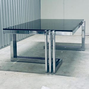 May include: A modern coffee table with a rectangular black glass top and a chrome-finished metal frame. The table has a geometric design with vertical supports on the sides. The table measures approximately 48 inches long, 24 inches wide, and 16 inches high.