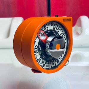 Può includere: Una radio National Panasonic AM/FM arancione con quadrante e antenna argentati. La radio ha un design circolare con un quadrante in bianco e nero. Il marchio è stampato in bianco. Un dispositivo elettronico vintage.