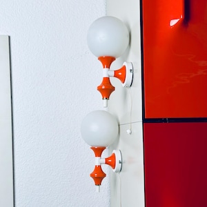 Könnte beinhalten: Zwei Wandleuchten mit kugelförmigen weißen Glasschirmen, die von leuchtend orangefarbenen und weißen Sockeln getragen werden. Jede Leuchte verfügt über eine kleine Zugschnur mit einer weißen Perle, montiert an einer weißen Eckfläche.