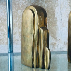 May include: A gold-colored, Art Deco-style sculpture with a vertical, layered design. The sculpture has a smooth, polished surface and is displayed on a glass shelf. The piece has a rectangular base and a rounded top.