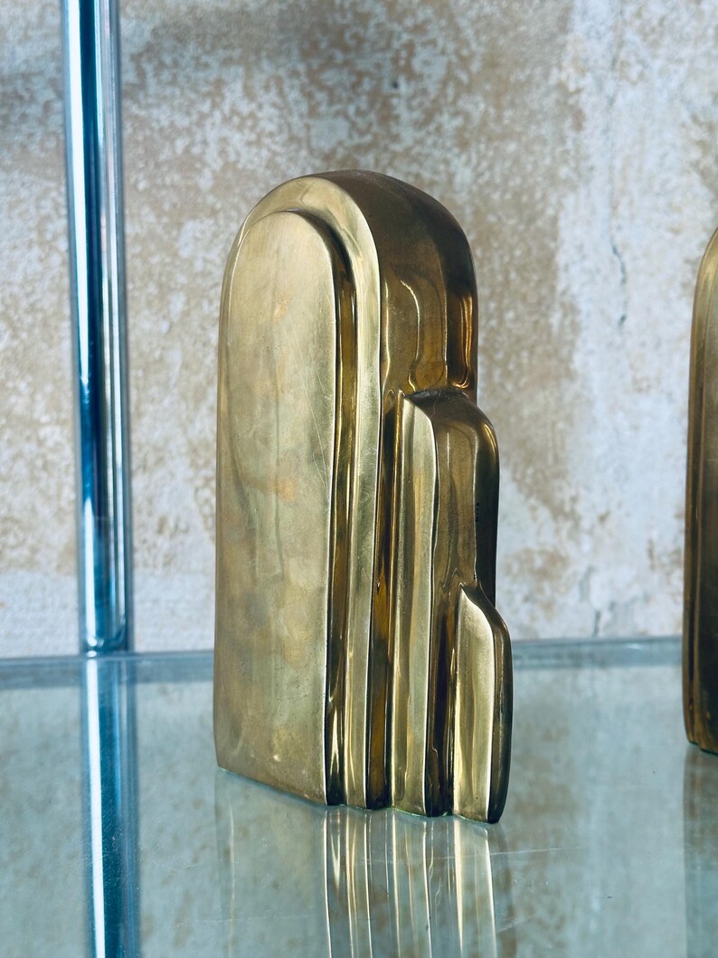 May include: A gold-colored, Art Deco-style sculpture with a vertical, layered design. The sculpture has a smooth, polished surface and is displayed on a glass shelf. The piece has a rectangular base and a rounded top.