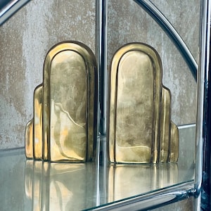 May include: A pair of Art Deco style bookends in a polished brass finish. The bookends have a stepped design with rounded tops and are displayed on a glass shelf. The background is a textured, neutral-toned wall.