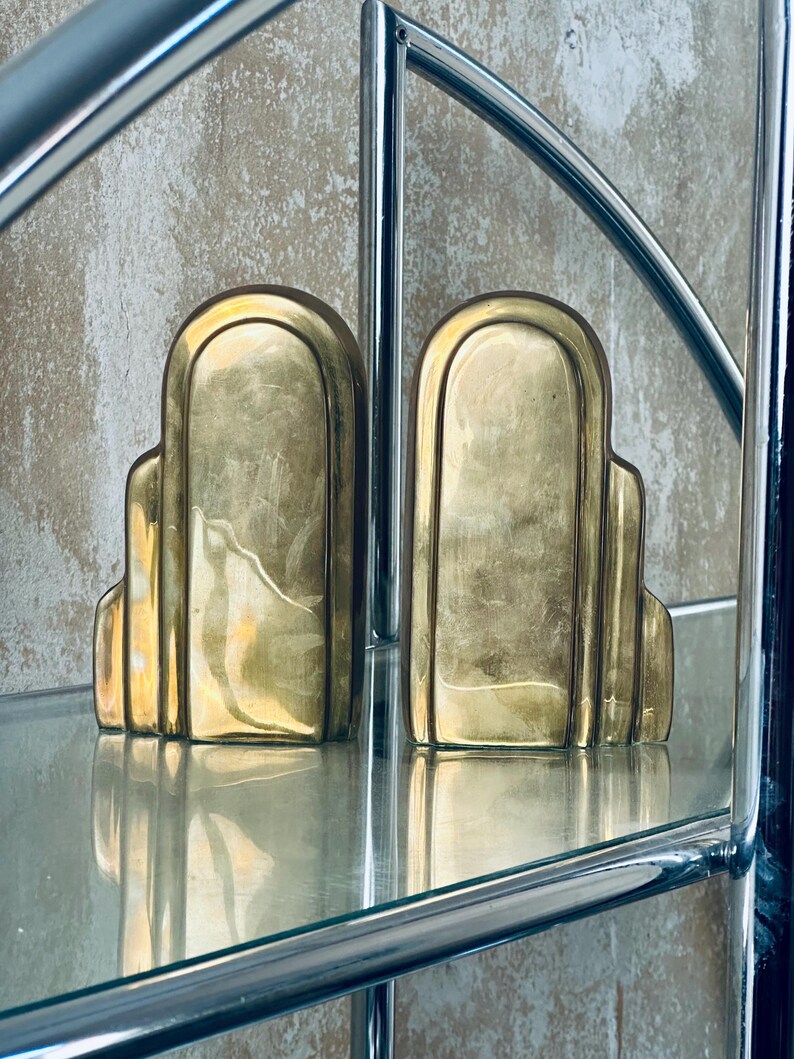 May include: A pair of Art Deco style bookends in a polished brass finish. The bookends have a stepped design with rounded tops and are displayed on a glass shelf. The background is a textured, neutral-toned wall.