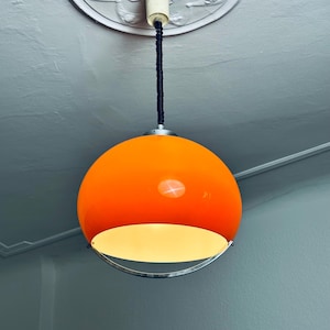 May include: A vibrant orange globe pendant light with a white and silver trim. The light fixture hangs from a black cord and is attached to a white ceiling fixture. The light is turned on, illuminating the room.