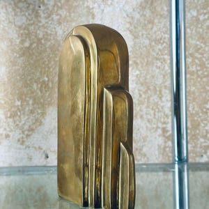 May include: A solid brass Art Deco style sculpture. The sculpture has a rectangular base and a rounded top with vertical lines and stepped details. The brass has a polished finish and reflects light.