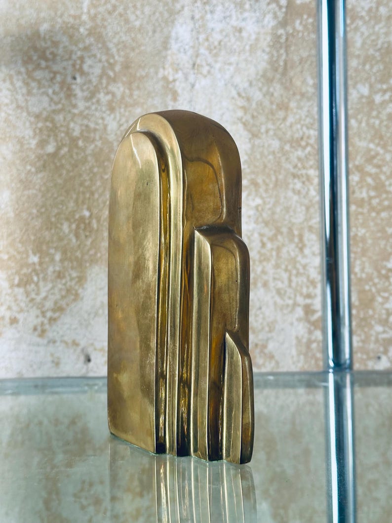 May include: A solid brass Art Deco style sculpture. The sculpture has a rectangular base and a rounded top with vertical lines and stepped details. The brass has a polished finish and reflects light.
