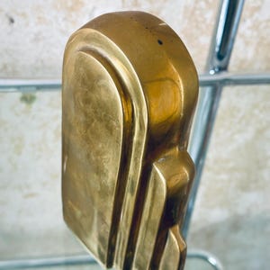 May include: A solid brass sculpture with a rounded top and a series of vertical, curved lines on the side. The sculpture has a smooth, polished finish and a warm, golden color. The object is displayed on a glass shelf.