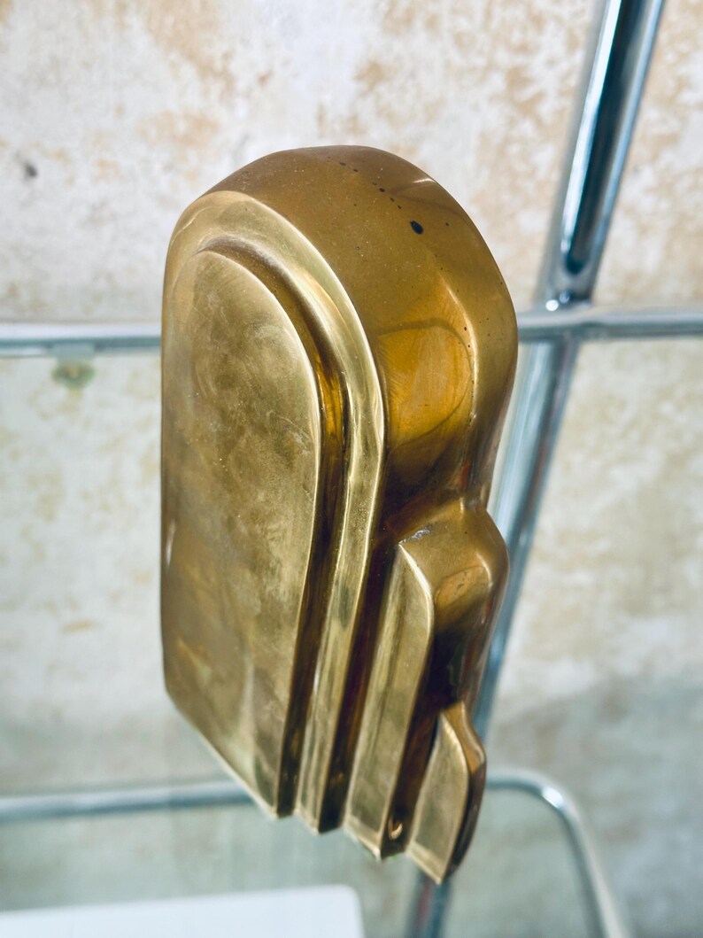 May include: A solid brass sculpture with a rounded top and a series of vertical, curved lines on the side. The sculpture has a smooth, polished finish and a warm, golden color. The object is displayed on a glass shelf.