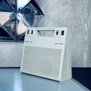 Może przedstawiać: Kremowy, zabytkowy Philips Radio Phono z uchwytem. Radio posiada kratkę głośnika z poziomymi liniami i pokrętłami. Widoczne jest logo Philips. Na boku widnieje napis "RADIO | PHONO".