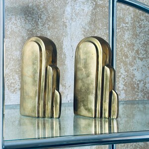May include: Two Art Deco style bookends in a gold finish. Each bookend has a rounded top and a stepped design. They are displayed on a glass shelf with a chrome frame, against a textured wall.