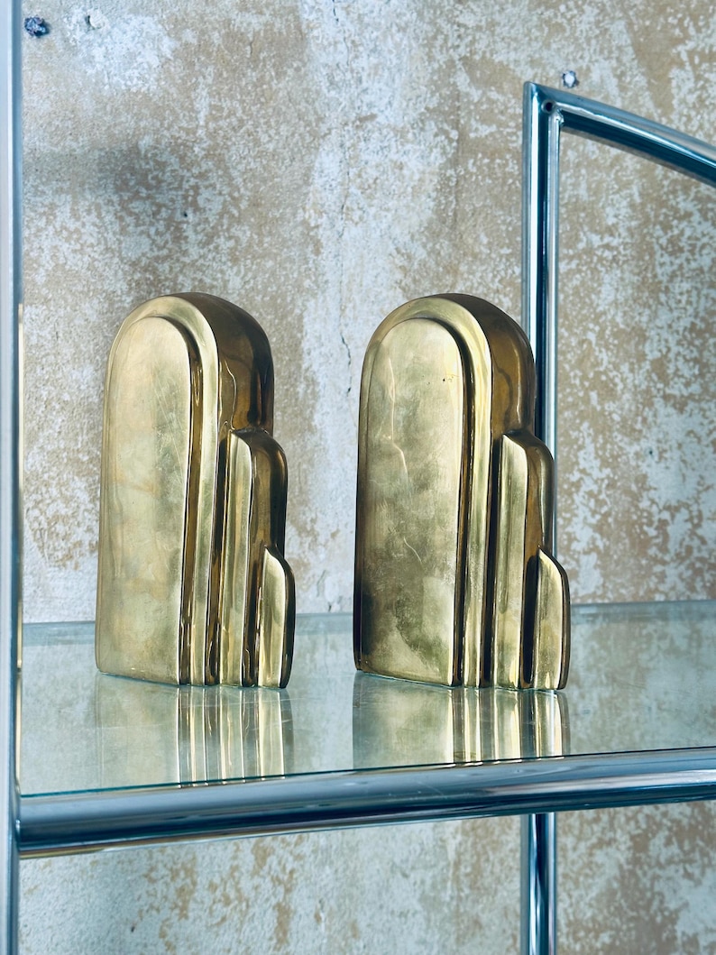 May include: Two Art Deco style bookends in a gold finish. Each bookend has a rounded top and a stepped design. They are displayed on a glass shelf with a chrome frame, against a textured wall.