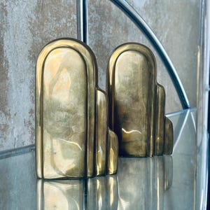 May include: A pair of Art Deco style bookends in a brass finish. The bookends have a stepped design with rounded tops and are displayed on a glass surface. The brass reflects light, creating a warm, vintage aesthetic.
