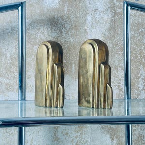 May include: Two Art Deco style bookends in a polished brass finish. The bookends have a stepped design and are displayed on a glass shelf. The background is a textured, neutral-toned wall. The bookends are a decorative home decor item.