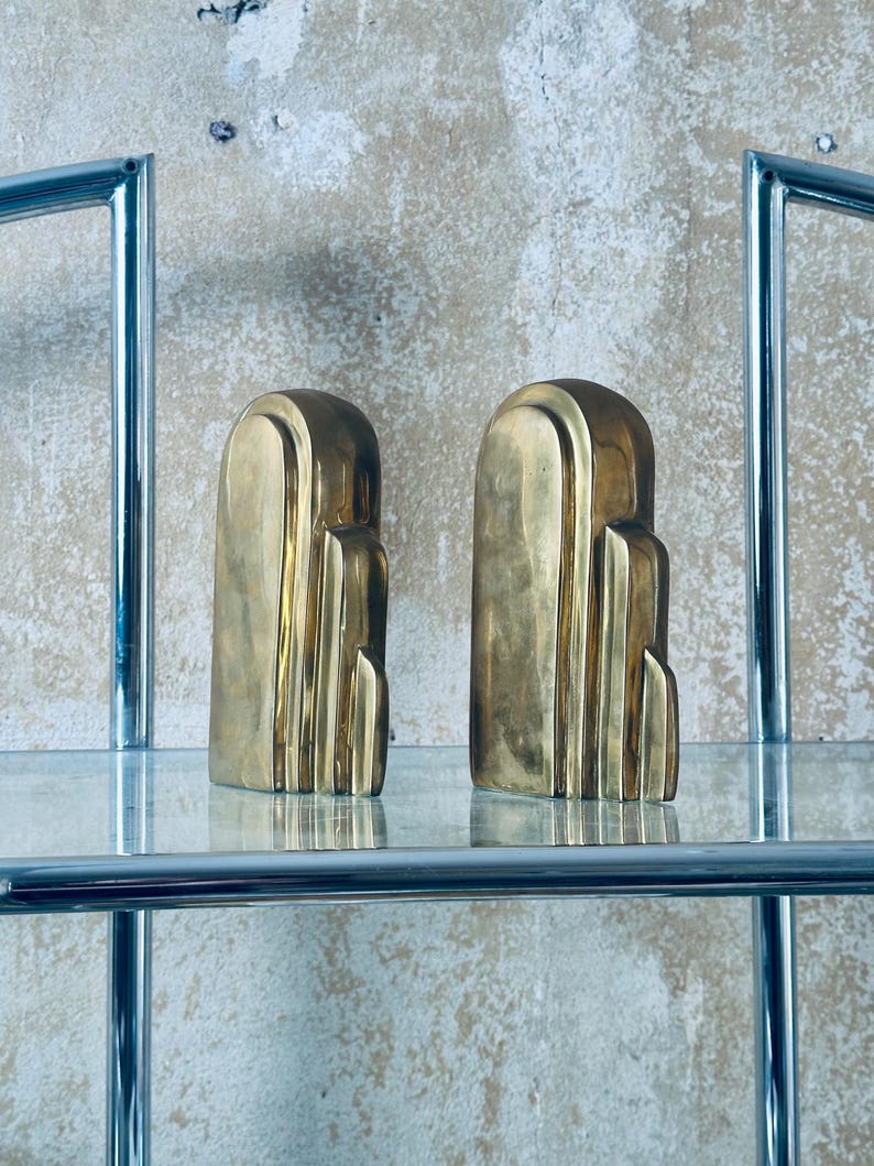 May include: Two Art Deco style bookends in a polished brass finish. The bookends have a stepped design and are displayed on a glass shelf. The background is a textured, neutral-toned wall. The bookends are a decorative home decor item.