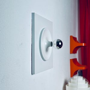 Könnte beinhalten: Eine weiße, quadratische Wandleuchte mit einem kreisförmigen Mittelelement und einer einzelnen hervorstehenden Glühbirne mit reflektierender, verchromter Spitze. Die Leuchte ist an einer flachen weißen Wand montiert und zeigt ein geometrisches Design.