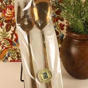 May include: A pair of gold-toned metal salad servers, consisting of a fork and a spoon, sealed in clear plastic packaging. A circular sticker on the front displays the text House of Siam, 8 Sathorn Nua, Bangkok, Thailand, and the logo HoS.