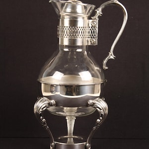 May include: A clear glass and silver coffee maker with a decorative handle and ornate base. The coffee maker has a bulbous glass body and a silver lid. The base has three curved legs. The coffee maker is designed to sit on a flat surface.