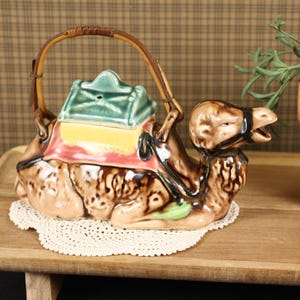 May include: A ceramic camel teapot with a brown and tan body, a green and yellow lid, and a woven handle. The teapot sits on a white lace doily on a wooden tray. A potted plant is in the background.