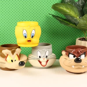 May include: Four ceramic mugs shaped like Looney Tunes characters. One yellow Tweety Bird mug is stacked atop a gray Bugs Bunny mug. To the left is a brown Wile E. Coyote mug, and to the right is a tan Tasmanian Devil mug with a wide grin.