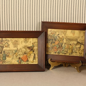 May include: Two framed vintage prints with dark wood frames. The prints depict scenes with figures in period clothing, possibly from the 18th or 19th century. One frame is propped on a gold-colored stand. A gold-colored decorative pot with a green plant is on the right.