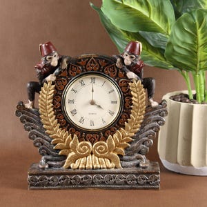 May include: A decorative table clock featuring two monkey figures wearing red fezzes, positioned on either side of a circular clock face with Roman numerals. The clock is framed by a gold-colored laurel wreath and intricate dark brown carved details.