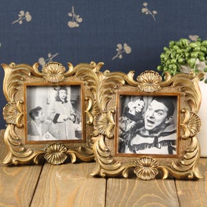 May include: Two square picture frames with ornate gold-toned borders featuring shell and scroll motifs. Each frame holds a black and white photograph. The left frame shows two people, while the right frame displays a person with a puppet character.