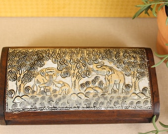 May include: A rectangular wooden box with a detailed relief carving of elephants in a forest scene. The box has a dark brown frame and a light-colored, intricately carved top. The elephants are depicted in a naturalistic style.
