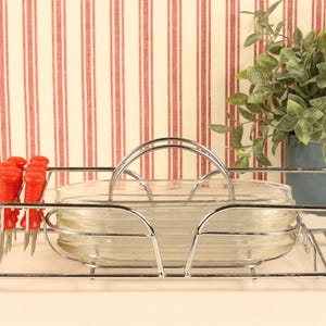 May include: A chrome wire serving caddy featuring a central handle and a rectangular base. The caddy holds a stack of clear glass oval dishes and a set of small metal appetizer forks with bright red plastic handles arranged on the side.
