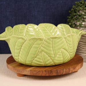 May include: A light green ceramic bowl with a leaf design, resting on a small, round wooden stand. The bowl's rim is shaped like leaves. A white vase with a textured pattern and greenery is in the background.