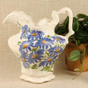 May include: A cream-colored ceramic pitcher featuring a painted pattern of blue daisy-like flowers with yellow centers and green leaves. The pitcher has a scalloped rim, a curved handle, and a molded relief design along the top edge and spout.