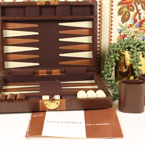 May include: A brown leather backgammon set with a gold clasp, open to reveal the playing board with brown, tan, and white triangles. Includes dice, checkers, and two brown leather dice cups. A booklet is in front.