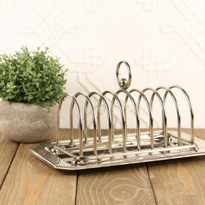 May include: A silver-toned metal toast rack featuring a rectangular base and a series of arched wire dividers. The rack includes a central circular handle at the top for carrying. The polished finish reflects light across its curved structure.
