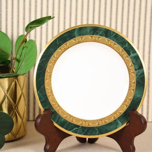 May include: A decorative plate with a white center, a green marble-like border, and gold accents. The plate is displayed on a wooden stand. A gold-colored pot with a plant is in the background.