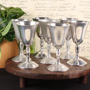 May include: A set of six silver-toned metal goblets with smooth, flared bowls and slender stems, each resting on a circular base. The goblets are arranged in a cluster on a round wooden platform, showcasing a polished, reflective metallic finish.