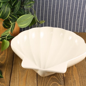 May include: A large, off-white ceramic bowl shaped like a scallop shell. The bowl features a smooth, glossy finish with deep, radiating ridges that extend from the base to the scalloped rim, creating a distinct, symmetrical, fan-like appearance.