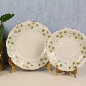 May include: Two white ceramic plates with a green floral design and gold trim, displayed on gold stands. A decorative gold pot with a plant is visible on the left side of the image. The plates are likely for serving food.