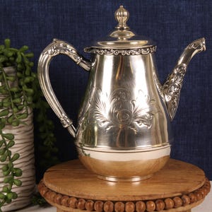 May include: A silver-colored teapot with ornate floral detailing and a decorative handle and spout. The teapot sits on a wooden stand with a beaded edge. A white vase with green plants is to the left of the teapot.