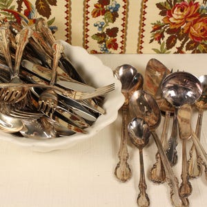 May include: A collection of antique silverware, including forks, spoons, and knives, displayed in a white, scalloped-edge bowl. The silverware has ornate handles and a shiny, reflective surface. A variety of serving spoons are also visible.