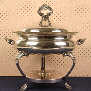 May include: A silver-plated chafing dish with a lid and ornate handles. The dish sits on a decorative stand with curved legs and a circular base. The lid has a decorative handle. The background is a dotted pattern.