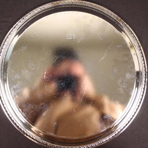 May include: A round, silver-colored serving tray with a decorative beaded edge. The tray's surface reflects light, showing a distorted reflection. The tray is set against a dark background, highlighting its bright finish.